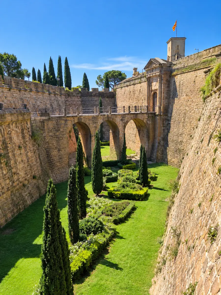 El foso ajardinado y cubierto de césped del castillo de Montjuïc entre murallas de piedra en talud, con un puente de arcos que lleva a la puerta