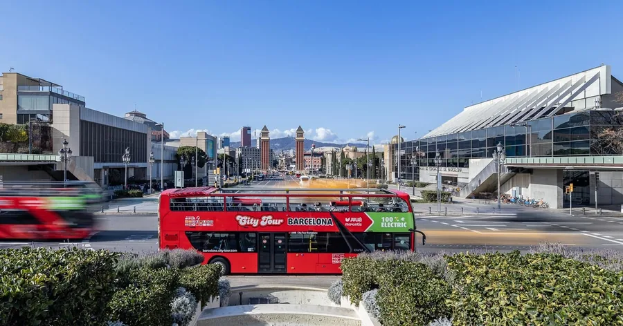 Un autobús turístico rojo en la plaza de España, con los antiguos pabellones de la Exposición detrás