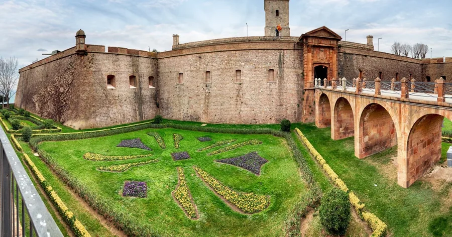 El castillo de Montjuïc desde arriba: gruesas murallas de piedra, un escudo de parterres en el foso y el puente de arcos de la entrada